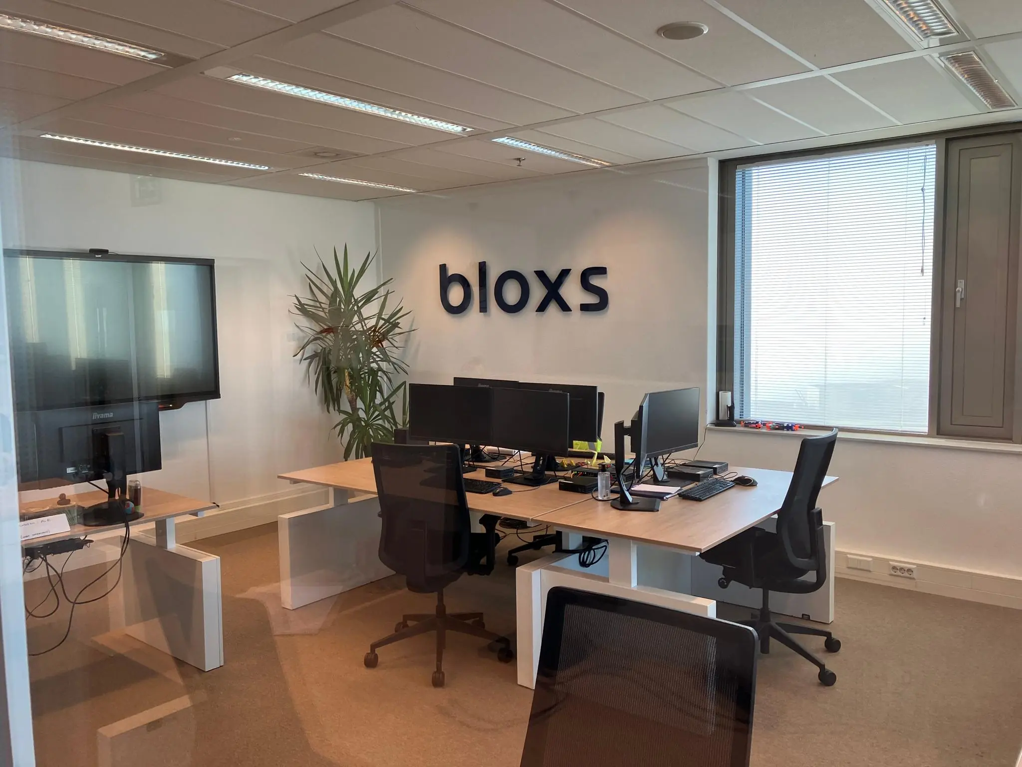 Modern kantoor van bloxs met werkplekken, computers en een groot raam met gesloten jaloezieën.